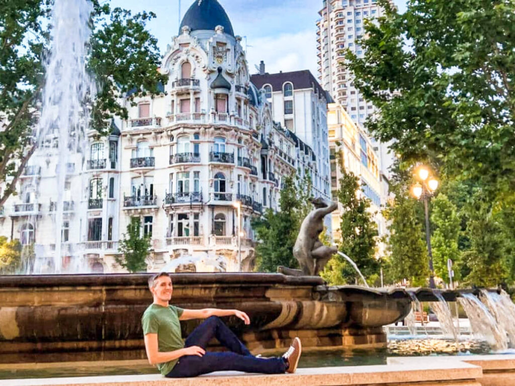 Nate Hake sitting near the fountain with the beautiful structures in the background in Spain